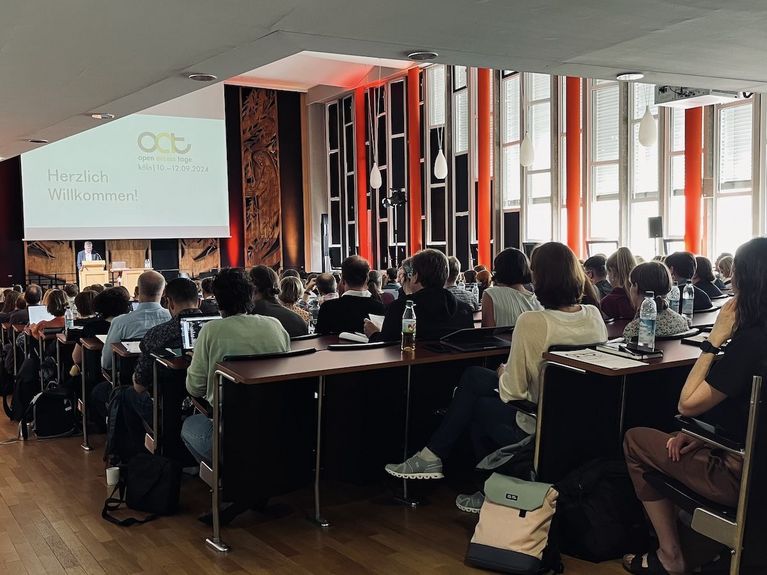 View into a lecture hall with fully occupied rows of chairs and a podium where a person is standing at a lectern in front of a presentation slide with the logo of the Open-Access-Tage 2024 and the text “Welcome!”